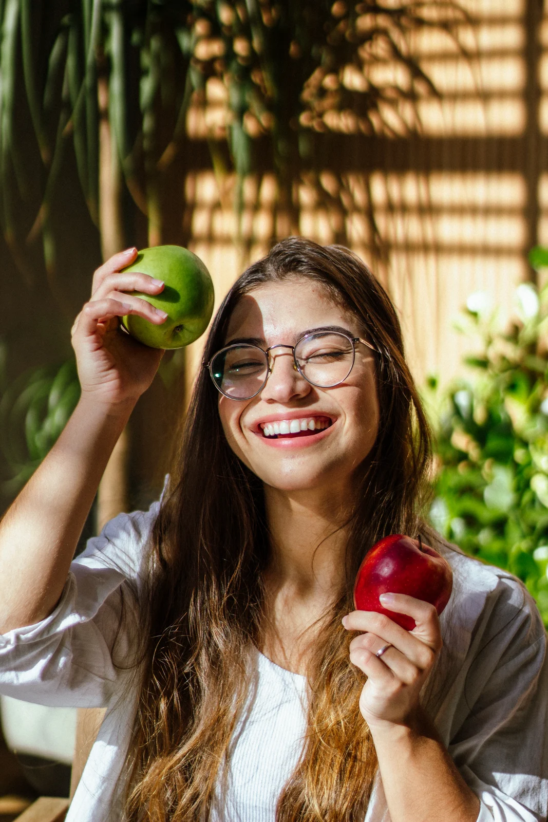 Duda sorrindo e segurando maçãs, em meio a plantas e luz natural